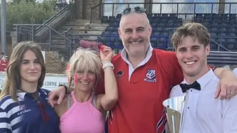 Family photo A woman with dark hair wearing a rugby shirt, a woman with blonde hair and wearing sunglasses on her head, a short-haired man wearing a red rugby shirt and a young man wearing a white shirt and a bow tie and holding a trophy, smiling at the camera with their arms around each other. They appear to be in a sports stadium with rows of seats behind them.