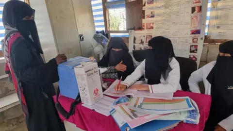 Three women sit behind a table, and another woman stands in front of it. There are paper and boxes on the table and posters on the wall.