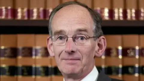 Royal Courts Of Justice A man with short grey hair and glasses looks at the camera with a neutral expression. He is facing the camera with shelves of books behind him.