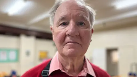 Emma Baugh/BBC Rex Birchenough who has short white hair. He is wearing a pink, collared shirt under a red jumper and is standing in a community-hall type building.