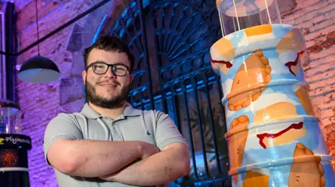 St Luke's Hospice Plymouth/Paul Slater Images Dafyd has short dark hair and a beard. He is standing with arms folded next to a lighthouse sculpture painted with pasties, ice cream and scones against a blue sky with fluffy white clouds