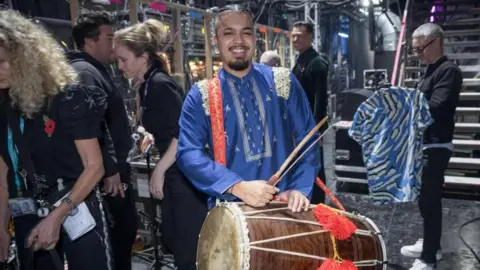 Strictly Come Dancing A man standing backstage at a television studio. He is wearing a blue outfit with gold embroidery. He has dark hair that is in two tight plaits. He is holding a large wooden drum with red pom poms. Television crew are in the background, asll wearing black clothes. 