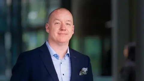 Professor Ken Sloan is seen in an official photograph. He is wearing a light blue shirt and navy jacket and the background is blurred.