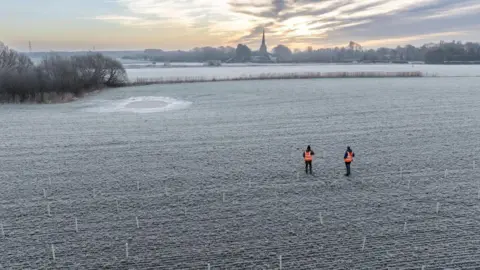 Paul Harris/National Trust Two men in hi-vis in a field frosted with ice where trees are being planted in Lunt with a church spire in the background