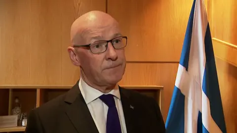 John Swinney is solemn-faced as he looks at the camera, the room has wooden panelling and a Saltire flag