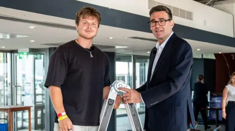 Andy Burnham, in a suit jacket and open necked shirt, stands at the top of a step ladder in an open plan office, opposite Max Klymenko, a young man with a light beard, wearing a black T-shirt