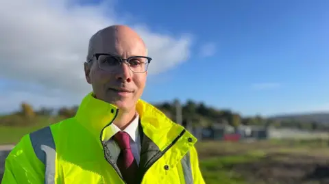 Peter sadler wearing glasses, a hi-vis jacket and a white shirt and maroon tie with a blurred countryside background