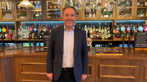 BBC Pub boss Nick Mackenzie, dressed in a casual suit, stands in front of one of his Greene King pubs. The wood-panelled bar has a selection of beers on draught, a fully stocked bar in the background also has some Halloween decorations