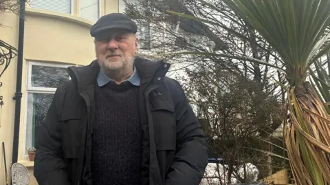 Alexander Hoare is standing in a garden with a palm tree on his left. He is smiling and wearing a flat cap, black anorak and blue jumper over a denim shirt. He has a white beard.