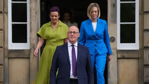 John Swinney wearing a dark suit and purple tie standing on the steps of Bute House. Behind him, members of his cabinet are emerging from a door. 