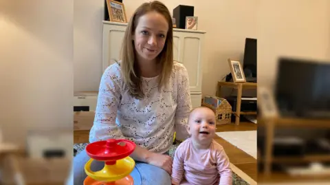 BBC Alice Cook wearing a white blouse and sitting on her living room floor beside her baby, Henrietta, who is wearing a pink stripey baby grow and smiling at the camera. 