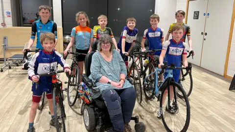 Six children in a assorted sports lycra stand holding bikes, in front of them are two boys also in cycling lycra holding bikes either side of a lady with round glasses and grey hair. She is in a wheelchair and is wearing a teal top and navy trousers, her right knee has a knee brace on it. 