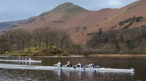 Clive Penkett Two rowing boats race across Derwentwater with mountains and a clear blue sky behind them. On an island you can just see a marshal wearing a hi-vis jacket 