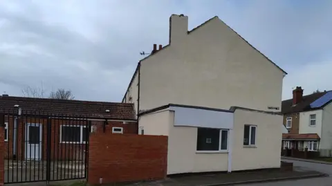A white building pictured side-on, with a redbrick extension behind it.