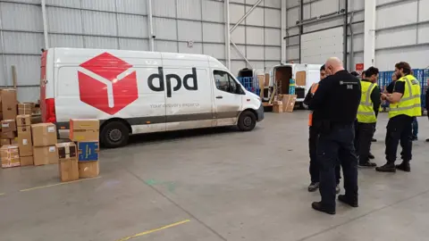 Men in high visibility jackets stood in a warehouse talking with a dpd van and parcels in the background.