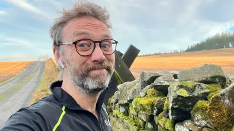 Matt Cole A man takes a selfie next to a stone wal. He is wearing a black top and has one earbud in. He has a beard and is wearing black glasses.