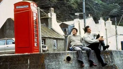 A shot from the movie Local Hero, two actors sitting at a harbour next to a red phone box.