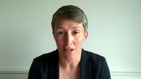 A woman with short hair and a dark top sits in a plane room. She is on a video call and is looking directly at the screen.