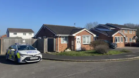 A police car parked outside a bungalow, with other houses alongside