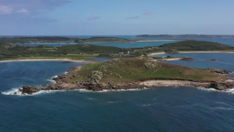 The island of Bryher is shown with rocky hills, sandy beaches and bodies of water beneath a blue sky scattered with clouds. 