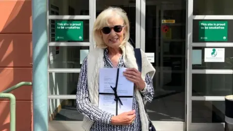 BBC Andrea Lewis outside a government building called Sir Charles Frossard House, holding a petition wrapped in a black ribbon. She has white hair and is wearing a pattered shirt and sunglasses, and has a white jumper over her shoulders.