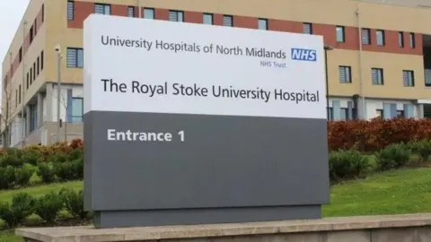 UHNM A white and grey sign outside The Royal Stoke University Hospital, with the hospital building visible behind it.