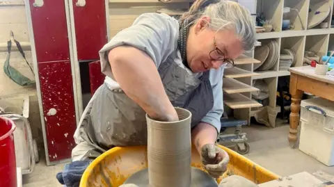 A woman is sat at a potter's wheel hand-making a tall clay pot. The has grey, tied-back hair, glasses, a grey apron and light blue shirt with rolled up sleeves. One of her hands is covered in clay beside the spinning pot, the other is inside the pot. The wheel has a yellow guard and the studio behind her has red lockers splattered with clay, white shelving, drying units and a working table.