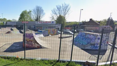 Google A skatepark, with fencing round it, showing graffiti on a large number of ramps. There is metal railing at the front of the photo and trees in the distance. 