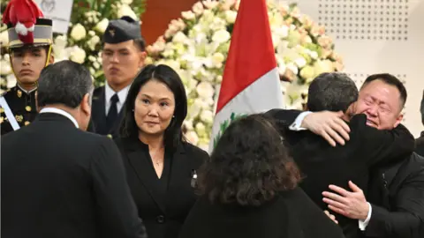 Getty Images Keiko dan Kenji Fujimori, anak-anak mendiang mantan presiden Peru Alberto Fujimori, menerima ucapan belasungkawa dari kerabatnya. Kenji, di sebelah kanan, terlihat memeluknya sambil menangis. Keiko di sebelah kiri dan tengah gambar menjabat tangan seorang pria yang membelakangi kamera.