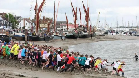 Chris Radburn/Fixedpoint Media A group of about 100 people run down the mud into the River Chelmer estuary. Boats on the mudflats and buildings are in the background. 