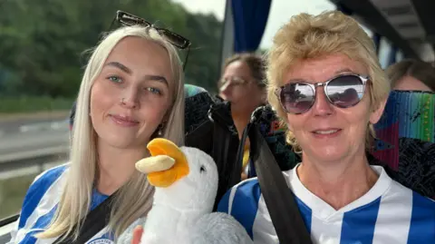 Amy Marchant and Sara Marchant sat in coach seats with a road visible out the window. They are holding a stuffed toy seagull with a chip in its mouth.