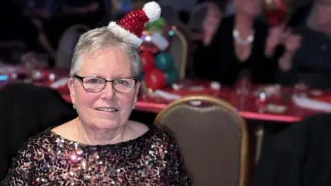 A woman wearing a sparkly party top and miniature Santa hat sits in a function room decorated for Christmas. She has short grey hair and wears glasses.