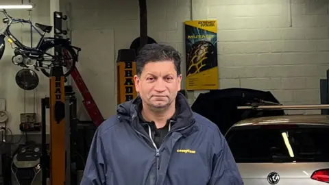 Zack Iqbal stands in the vehicle workshop of his business. A VW car is behind him, a bike is hanging up. He has short black hair and wears a blue rain jacket with black shirt underneath.