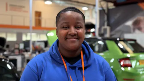 Olivia is wearing a bright blue hoodie and is behind cars.