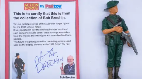 Vectis Auctions A 'Australian Jungle Fighter' figure of a man with a green hat and suit carrying a gun is displayed next to a certificate of authenticity featuring Bob Brechin's photograph and signature.