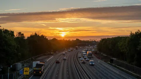 DereksDisco Two three-lane carriageways with several cars and lorries driving along are surrounded by a line of green trees on each side and overlooked by a low orange sun piercing through the clouds. 