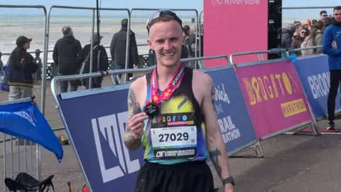 A man wearing a colourful running top and a a marathon number smiles while holding the medal around his neck. Several people walk along the seafront in the background.