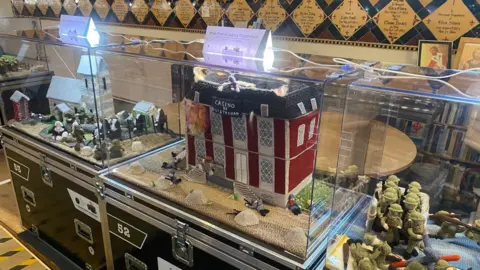 Three clear boxes, side by side, each contain an individual scene of war - with soldiers depicted one alongside a casino building in another and a rural village scene the other.