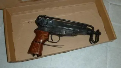 A black metal gun with a wooden handle in a cardboard box. The box is laying on top of white paper sheets. 