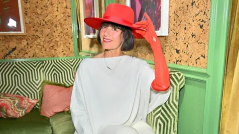 Getty Images Danielle is wearing a draped white dress and long, red gloves and a red hat. She is sitting down in a green, patterned room.