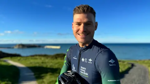 Sam Brand has short hair styled up. He is wearing a dark blue and green cycling top, smiling, and holding his helmet, standing on a headland, where the blurred outline of Peel Castle can be seen on his left, with the Irish Sea all around on a sunny day.