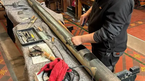 A large metal pipe, several metres long with open sections cut out, is lying on a table while someone works on it with a tool. The person's head is cropped out. Various other wooden handled tools are on the table.
