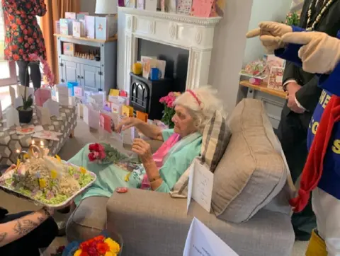 Edith surrounded by cards, people and being given a birthday cake