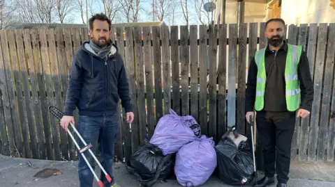 A man wearing a scarf and a dark hoodie and a man wearing a luminous green bib stand either side of some bags of rubbish. A wooden garden fence is in the background and they are both carrying litter picking tools.     