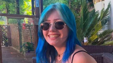 Morgan Dorsett, seen here with blue hair and sunglasses, sitting in what looks like an outdoor deck with palm trees and other greenery around.