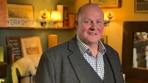 BBC/Olivia Richwald A bald man wearing a tweed suit looks at the camera