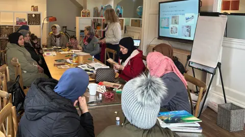 A group of women, mainly from Afghanistan are sat around a table looking at books.