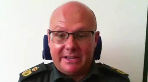A man called Wayne Darch. He is bald and slightly sunburnt. He is wearing a green uniform worn by ambulance workers. He has glasses on.