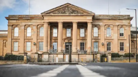 BBC The High Court in Dundee, a brown brick building with pillars at the front, under a cloudy blue sky.