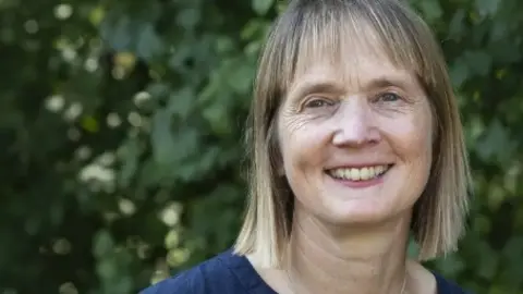 Suffolk Wildlife Trust Christine Luxton smiles at the camera while standing in front of a large bush. She has shoulder length blonde hair and a fringe. She wears a navy top.